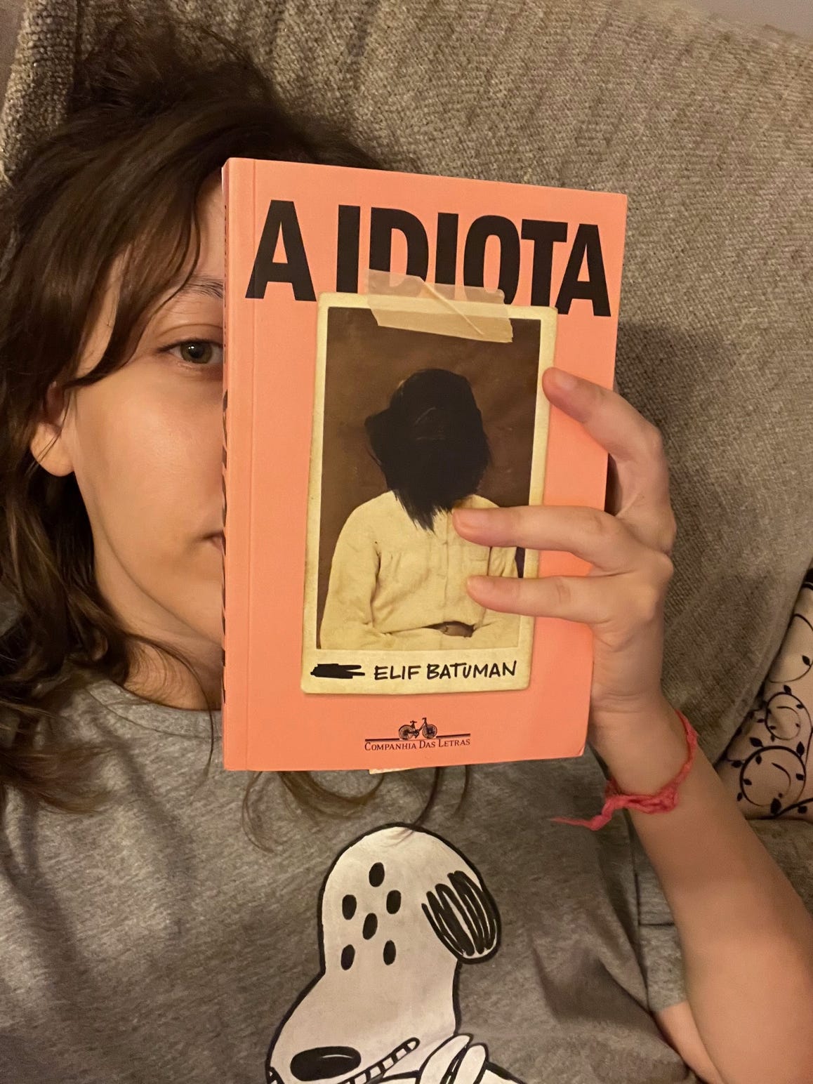 Selfie em que estou segurando a capa do livro no rosto, tampando a metade da face. Estou usando uma camiseta do Snoopy alienígena.