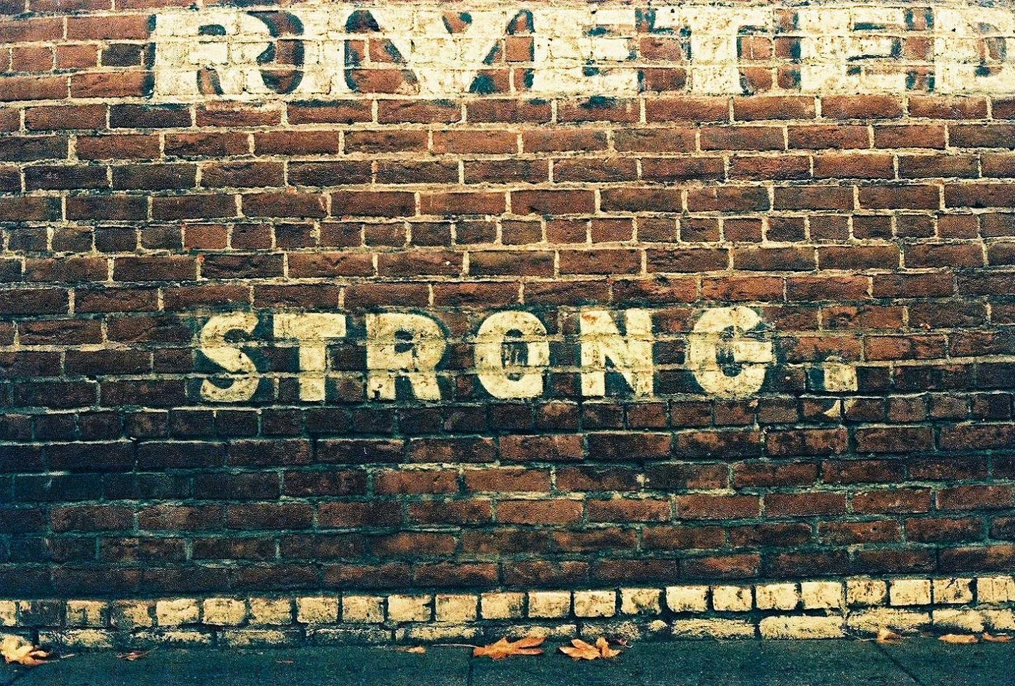 A brick wall with partial white lettering, which reads "RIVETED" near the top of the frame, and "STRONG," near the middle.