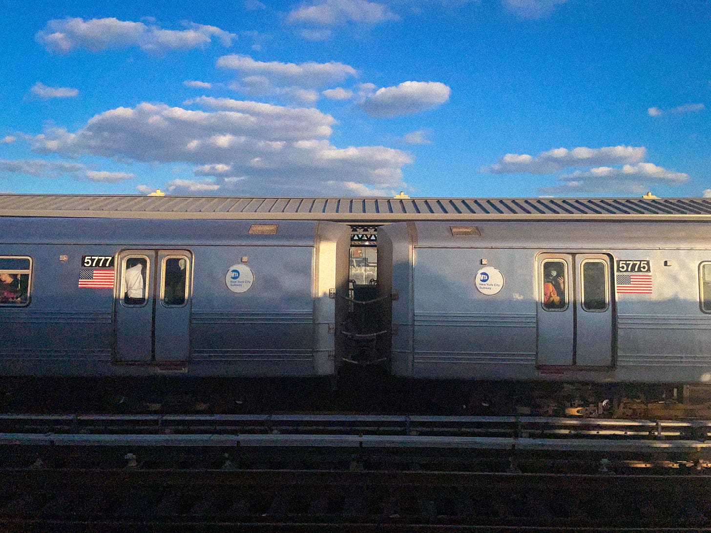 A NYC subway train under a blue sky dotted with clouds.
