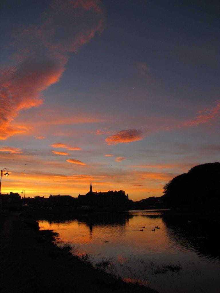 A beautiful sunrise, with the silhouetted outline of Wick, Caithness reflected in the river. Some ducks swim on the still surface and clouds are streaked with oranges and gold. Watching, or feeling, the first rays of the sun is always a rewarding part of inspiring travel. 