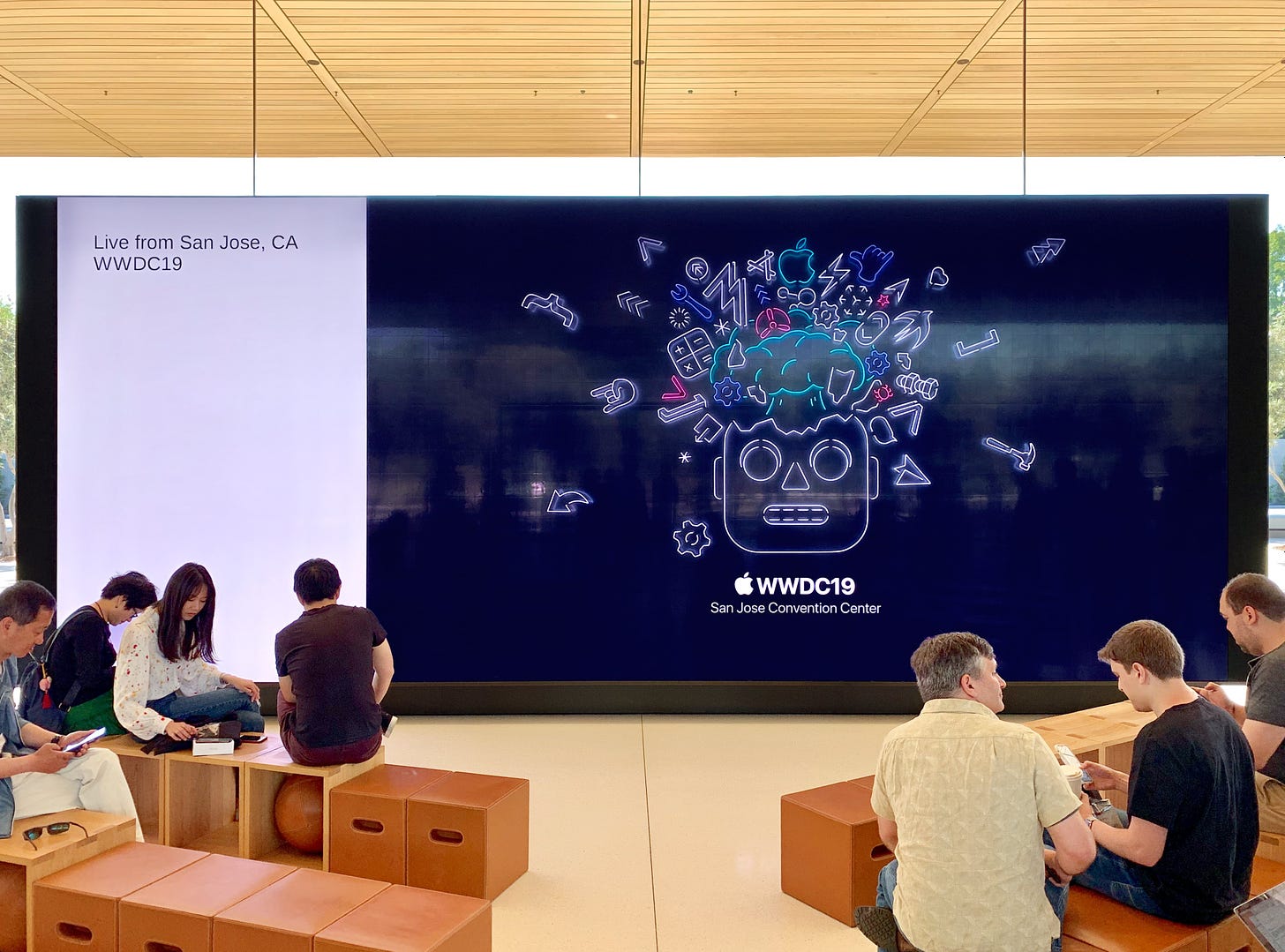 The Forum at Apple Park Visitor Center. On the video wall is artwork for WWDC 2019. People are sitting on Forum seats in front of the video wall, waiting for a livestream to begin.