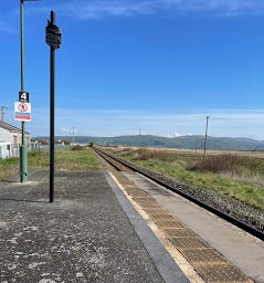 A shot of a train disappearing in the distance on a single rail track in a rural Welsh station. There are hills in the distance and fields either side of the track.