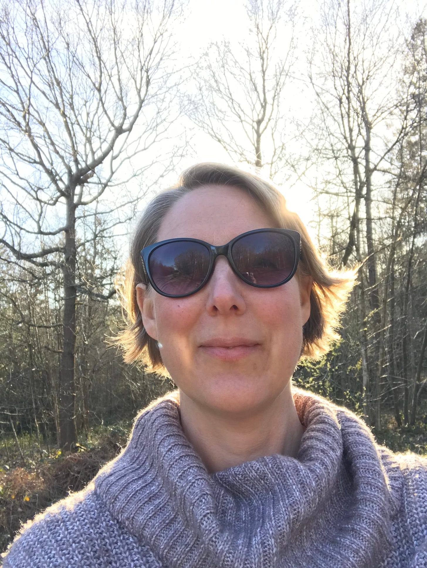 a photo of me, a white woman in sunglasses with short blonde hair smiling at the camera, I'm wearing a large brown jumper and standing in front of bare trees in the woods.