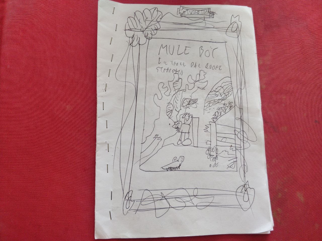 Sfondo rosso e foglio con un disegno da bambino che riproduce la copertina del fumetto "Mule Boy e il troll dal cuore strappato".