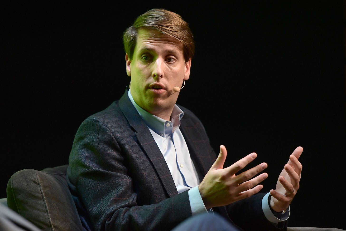 A white man in a dark suite jacket sits in front of a black, blank background