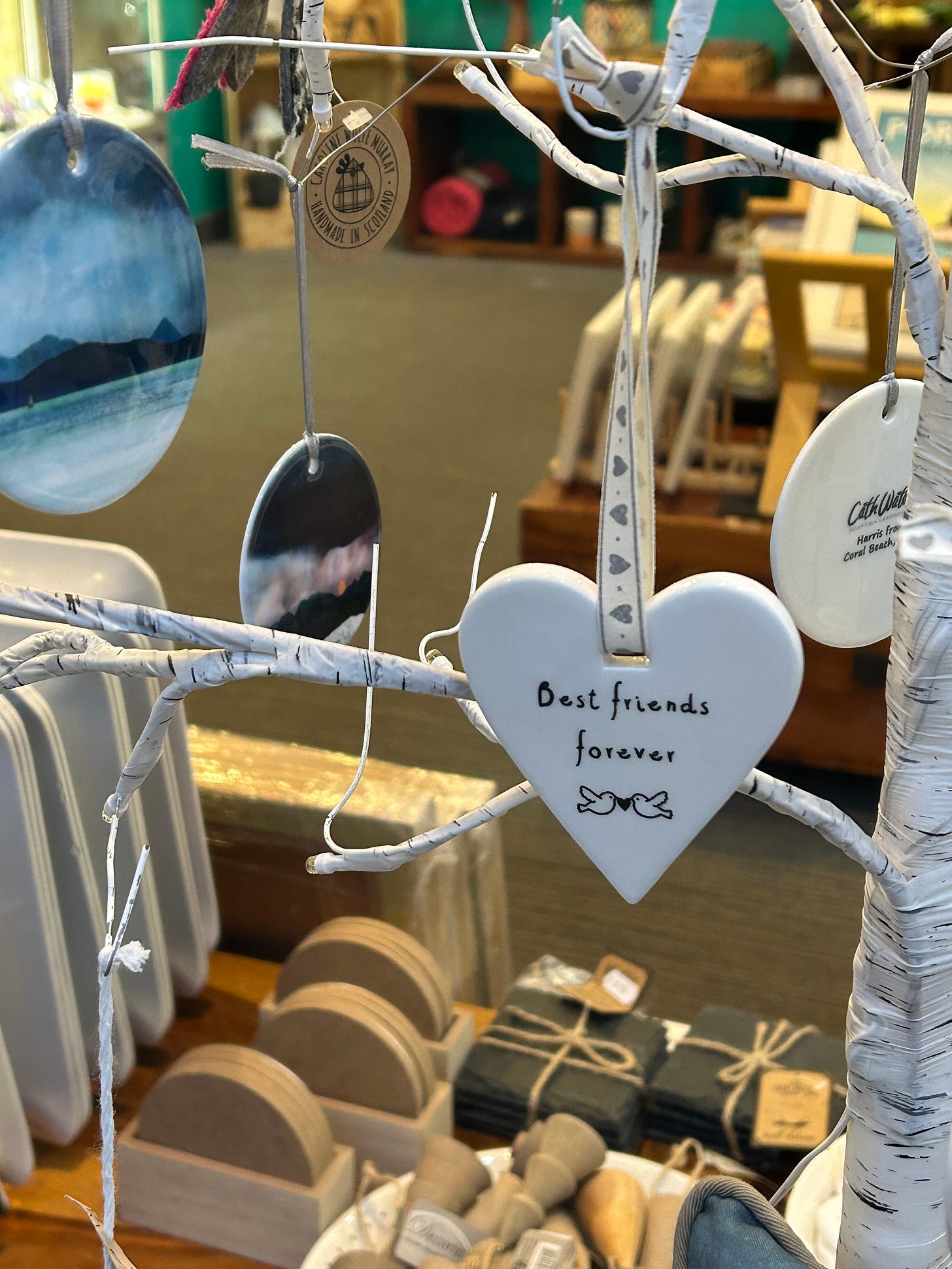 A storefront window display showcasing a white ceraminc heart hanging from a display tree. The heart has BEST FRIENDS FOREVER writen on it in black ink, above an illustration of two doves holding a heart between them, in their beaks.