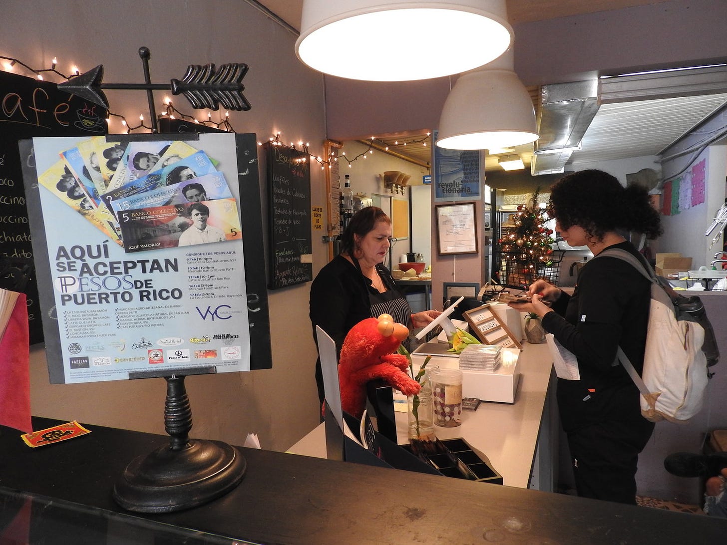 A cafe worker serving a customer with a sign on the counter explaining “Aquí se aceptan Pesos de Puerto Rico” (We accept Puerto Rico pesos) — the community currency of the Valor Y Cambio project.