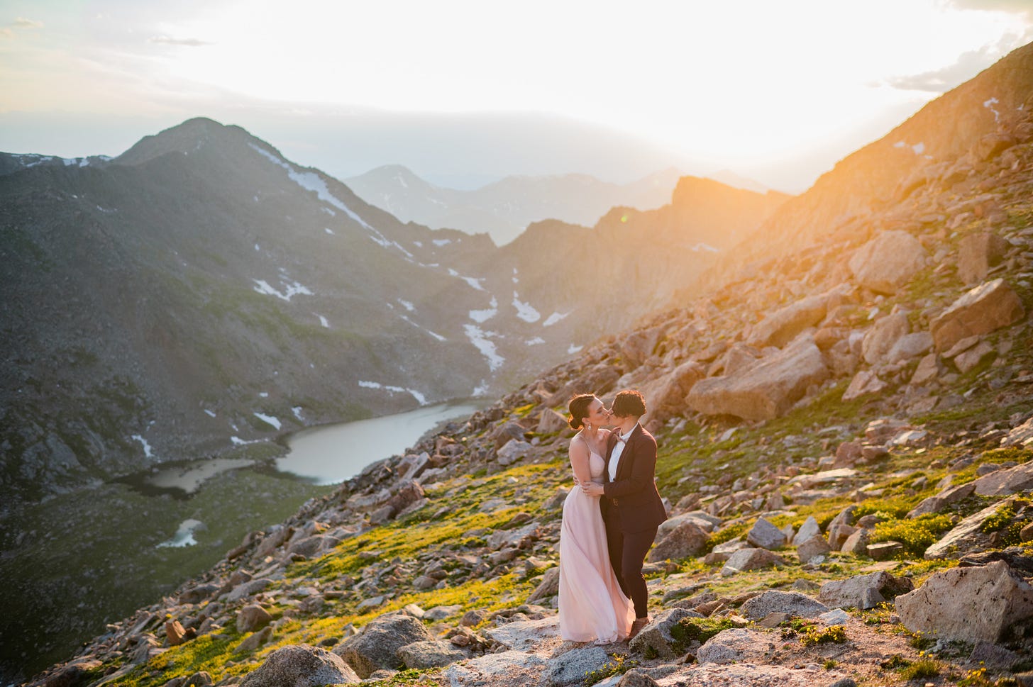 A photo of Shohreh and Jessie kissing in fancy clothes on the side of a mountain with miles of mountain rage and an alpine lake seen behind them in the distance