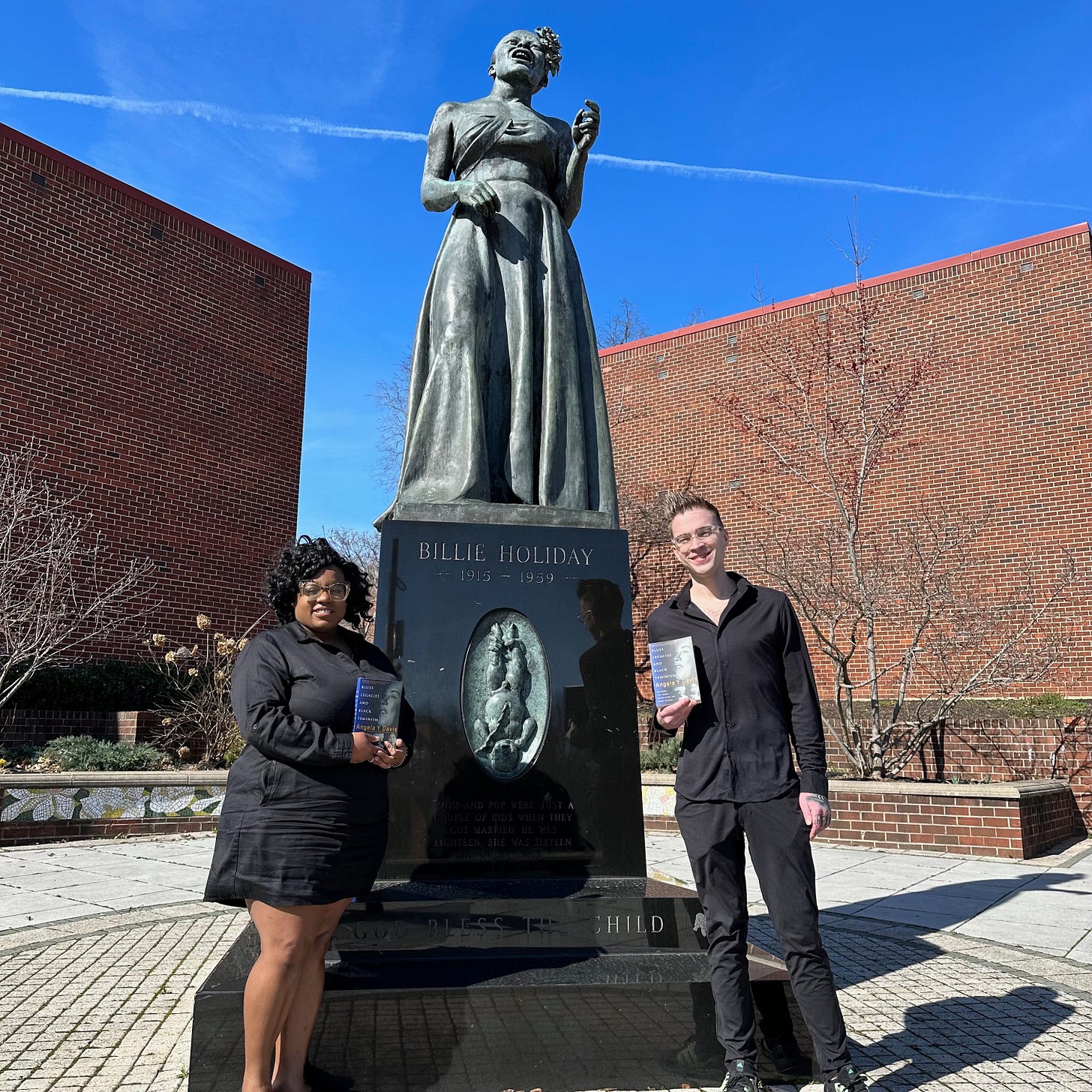 A color photograph of African American woman Tiheera Blount and white man Emmanuel Mehr posing beside the Billie Holiday statue in Baltimore. They both hold a copy of Angela Y. Davis's book Blues Legacies and Black Feminism, which features an image of Holiday on its cover.