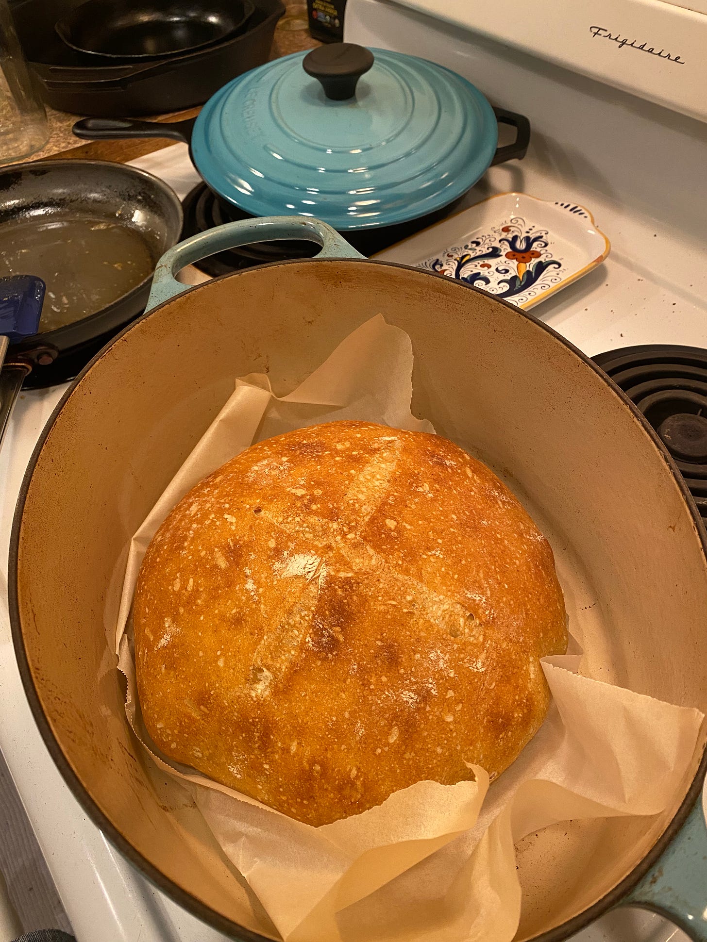 A sourdough boule with an X score on top, resting in an oval Le Creuset dutch oven with parchment on the bottom. The lid for the pot is sitting behind it on the stove.