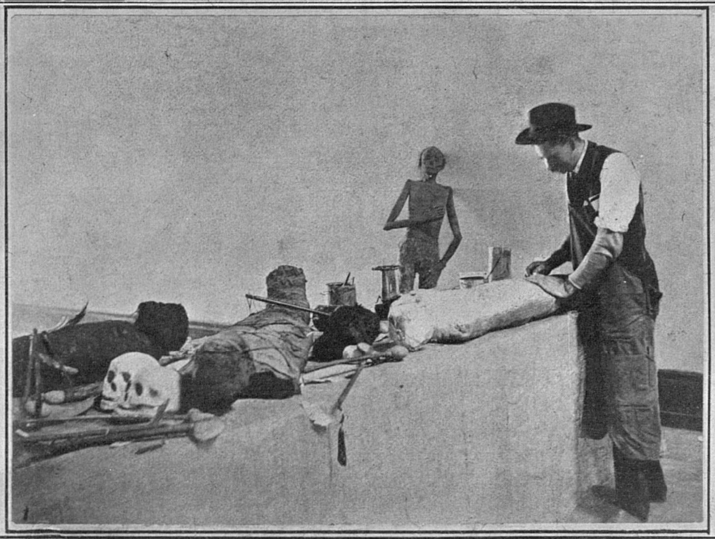 A man in early 20th-century American clothing stands at one end of a table where there are various unidentified objects. In the background, a human figure leans against a wall - its spindly arms and blank expression suggest it is a mummy.
