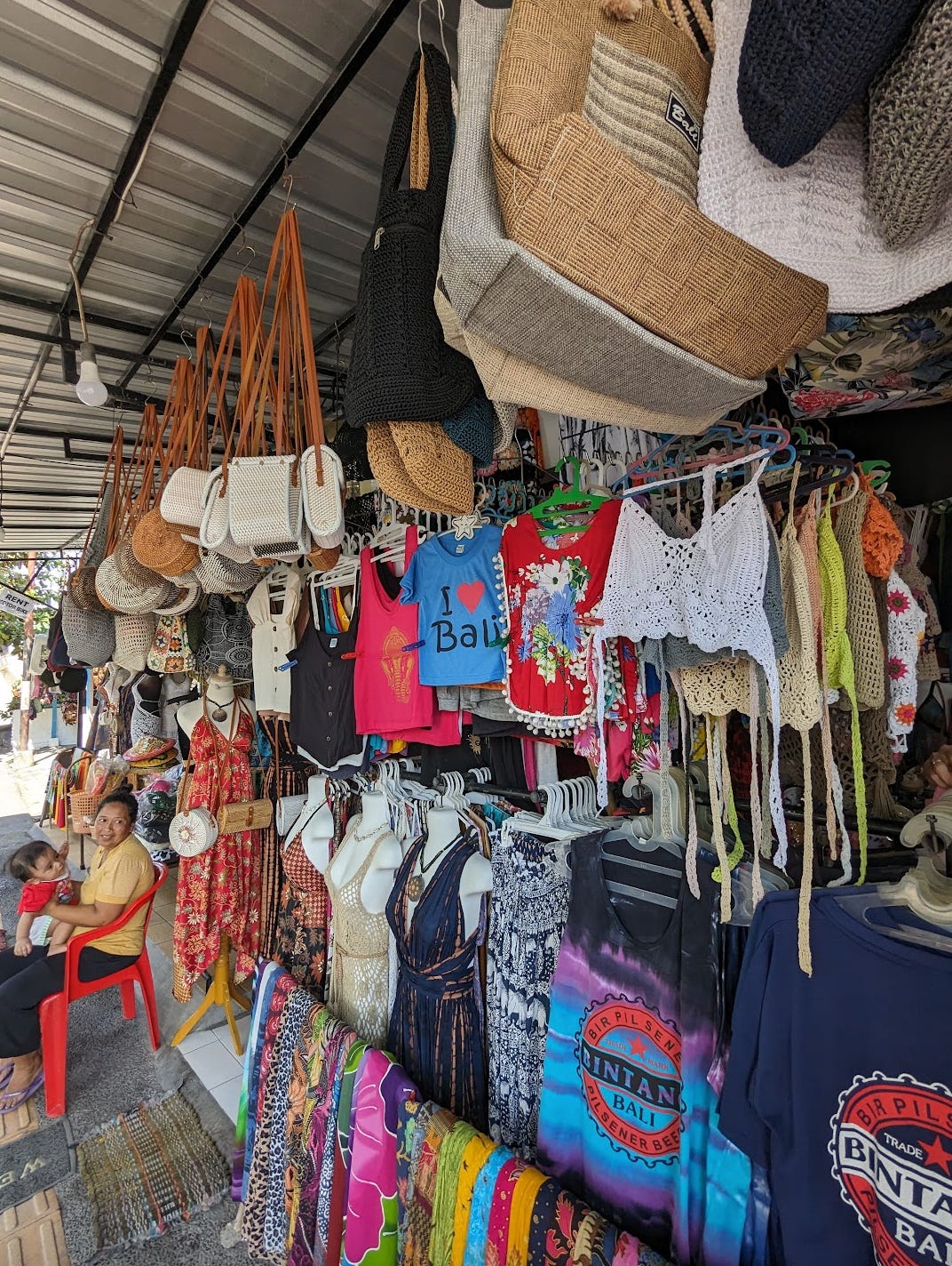 A shop with purses and clothes hanging from rods including a blue shirt that reads, "I <3 Bal". A woman sits on a red chair with a baby on her lap by the shop