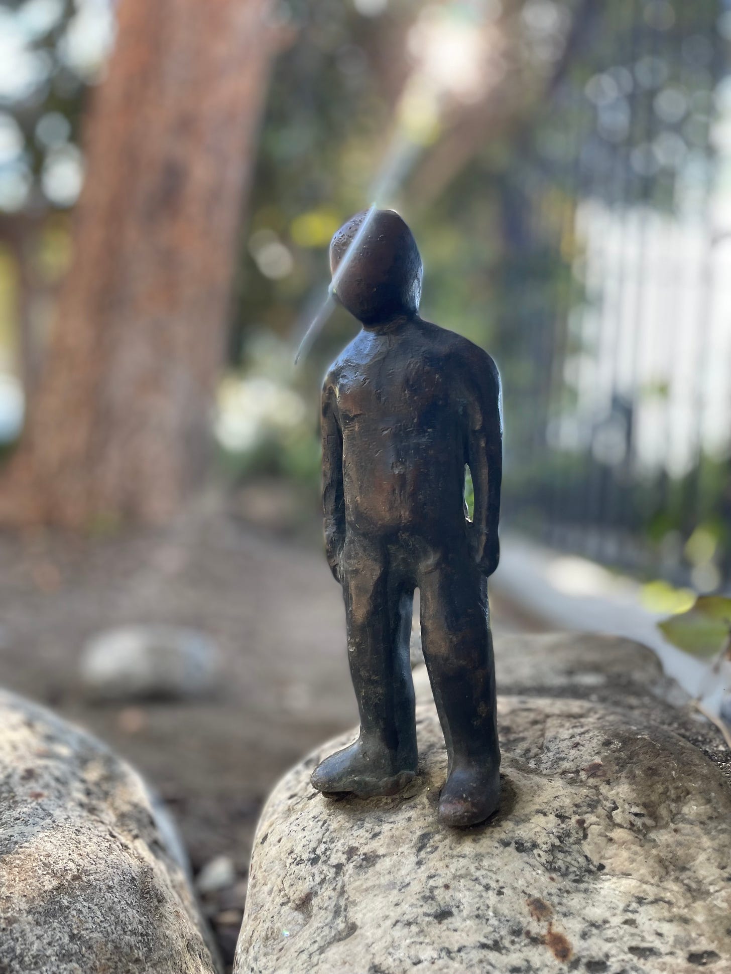 A picture of a small metal sculpture of a humanoid figure standing on a rock. There are blurred trees in the background. A small sunbeam is cutting across the statue's head from behind and shining through the front, like a burst of inspiration.