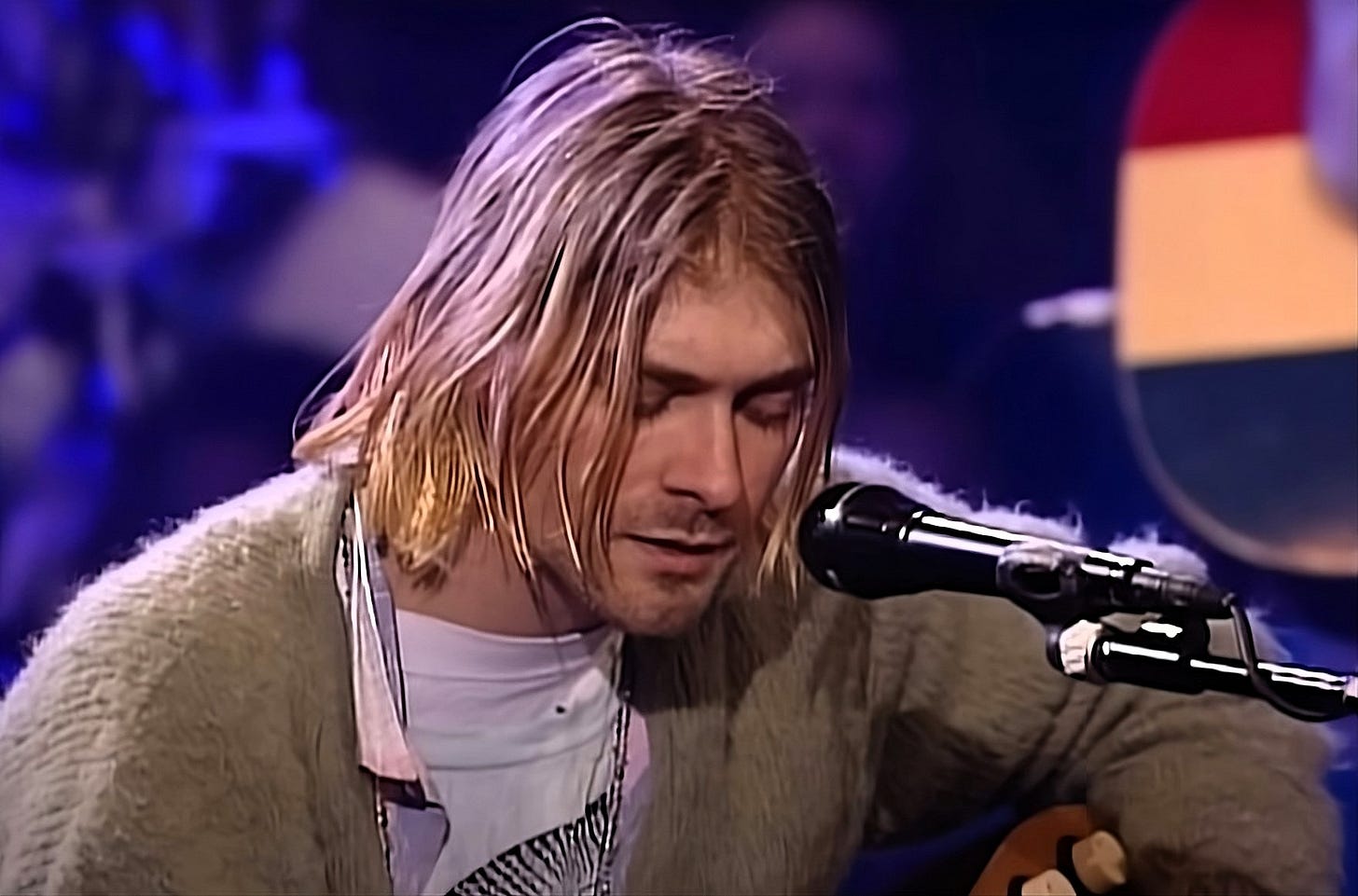 Kurt Cobain with long hair playing an acoustic guitar, singing Something in the Way into a microphone on stage. Kurt Cobain with long hair playing an acoustic guitar, singing Something in the Way into a microphone on stage.