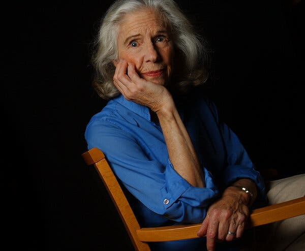 A close-up portrait of Ms. Sternhagen sitting on a wooden chair and turning to face the camera, her right elbow propped on one arm of the chair and holding her chin in her hand. She wears a royal blue shirt and has shoulder-length gray hair. 