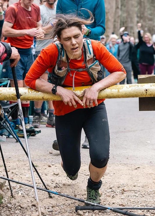 Jasmin Paris leans heavily on a yellow gate, signifying her historic finish of the 2024 Barkley Marathons. Her eyes are closed in a grimace and her hair is wild. Drool drips from her open mouth as she leans on the gate for support.
