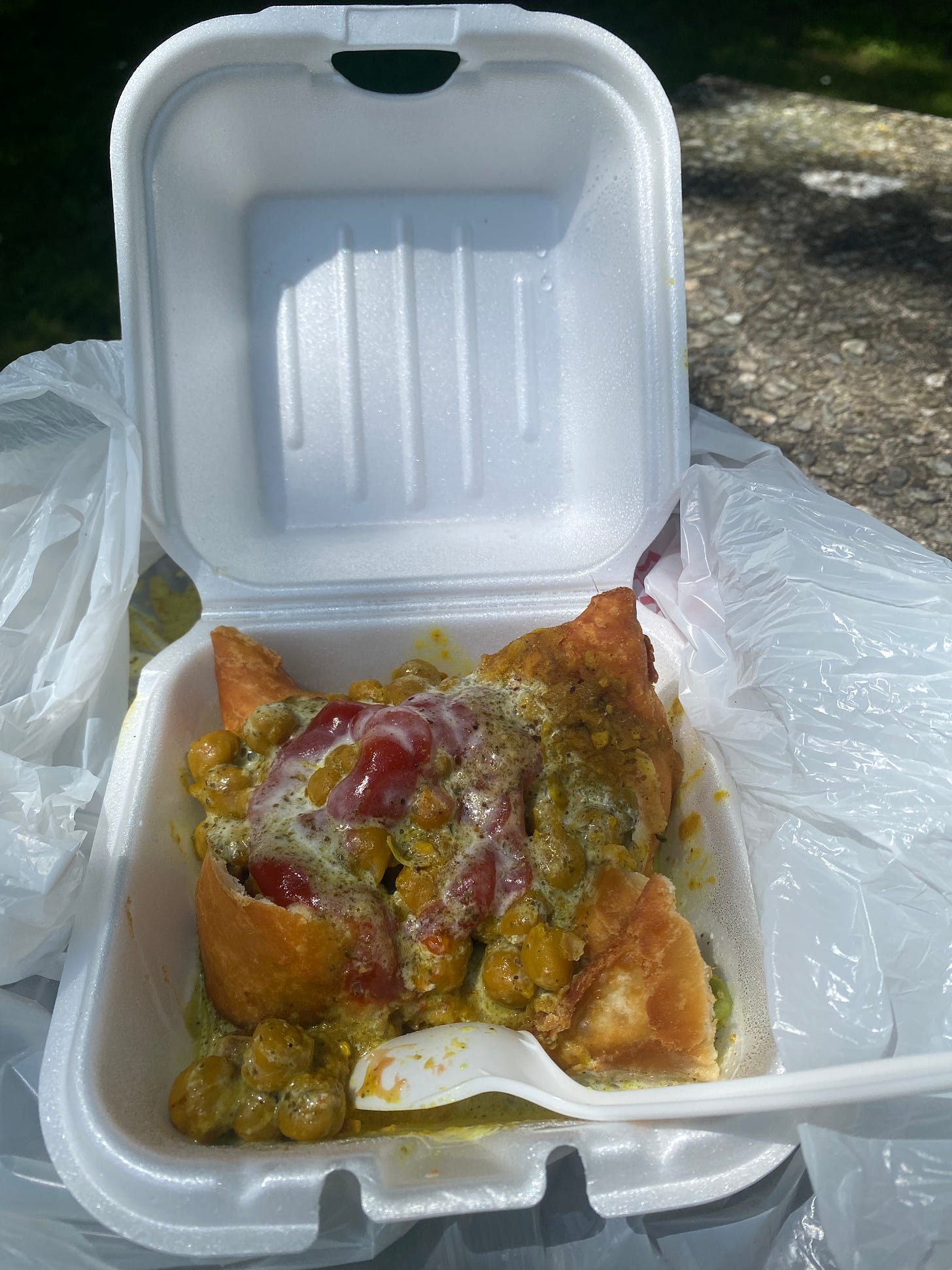 the samosa chaat described above: four corners of a samosa surrounded by chana curry and covered with ketchup and yogurt on top. It's in a small white takeout container with a plastic spoon sticking out of it.