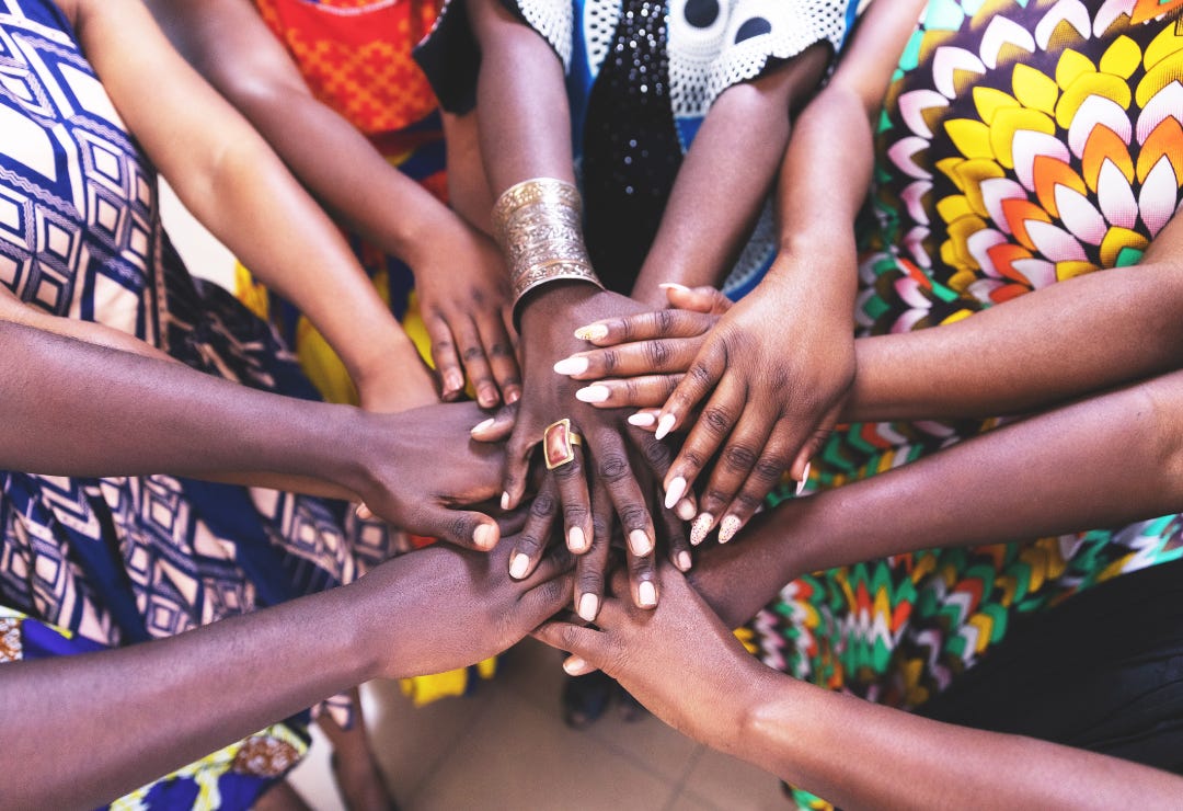 A group of colorfully-dressed African women gather their hands together in a community circle