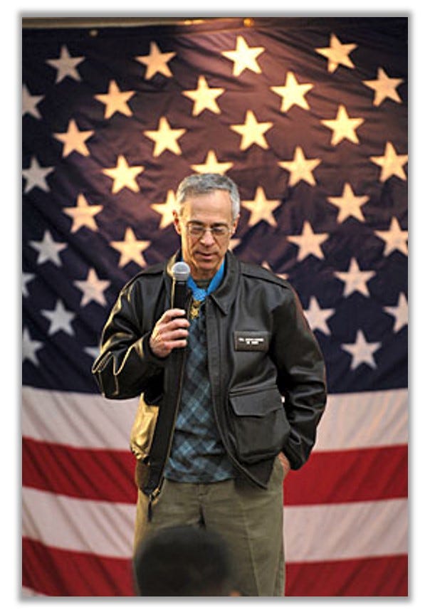 Jacobs is photographed addressing an audience. He is standing in front of an American flag backdrop.