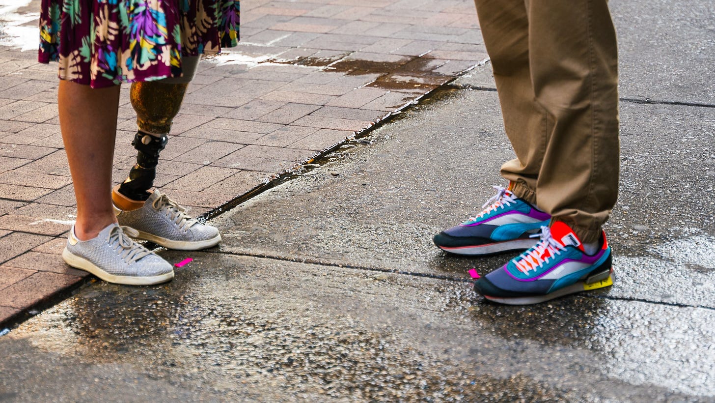 A shot of two people from the knees down on an urban sidewalk. On the right is a woman in a purple sundress with a colorful hawaiian-like design that goes below the knee. One of her legs is a prosthetic one. She's wearing blue tennis shoes. On the right is a figure in khaki pants with multi-colored shoes, including the colors purple, pink and violet blue with white laces. There's about two and a half between the two figures. A shot of two people from the knees down on an urban sidewalk. On the right is a woman in a purple sundress with a colorful hawaiian-like design that goes below the knee. One of her legs is a prosthetic one. She's wearing blue tennis shoes. On the right is a figure in khaki pants with multi-colored shoes, including the colors purple, pink and violet blue with white laces. There's about two and a half between the two figures.