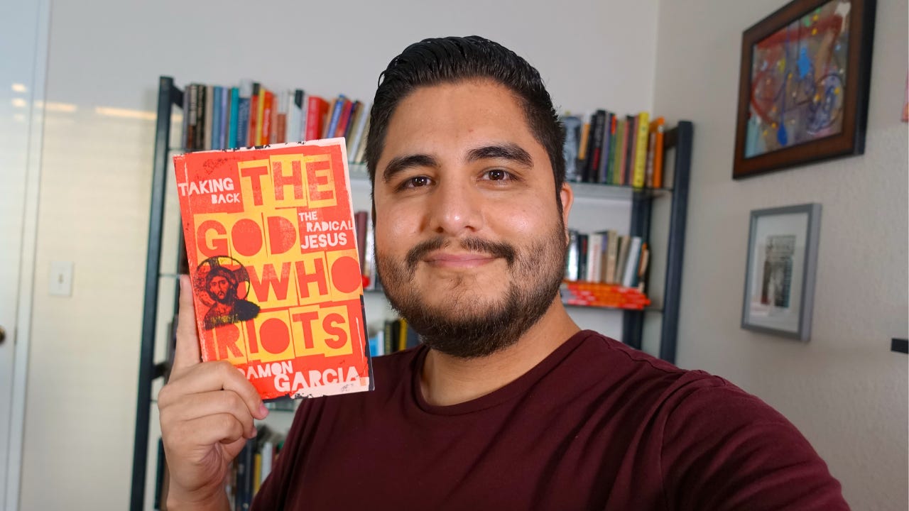Image of me smiling and holding up my book, The God Who Riots.