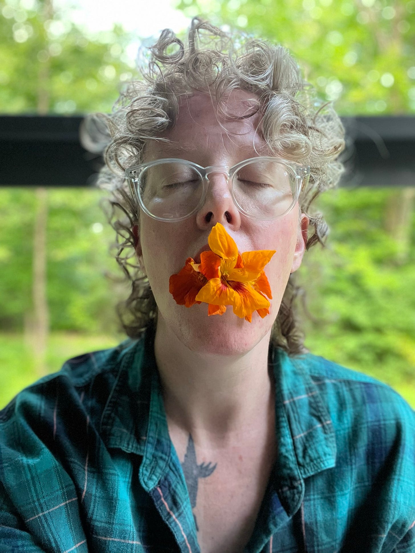 close portrait of a curly-haired, glasses-wearing person with their eyes closed, orange nasturtium blossoms coming out of their mouth