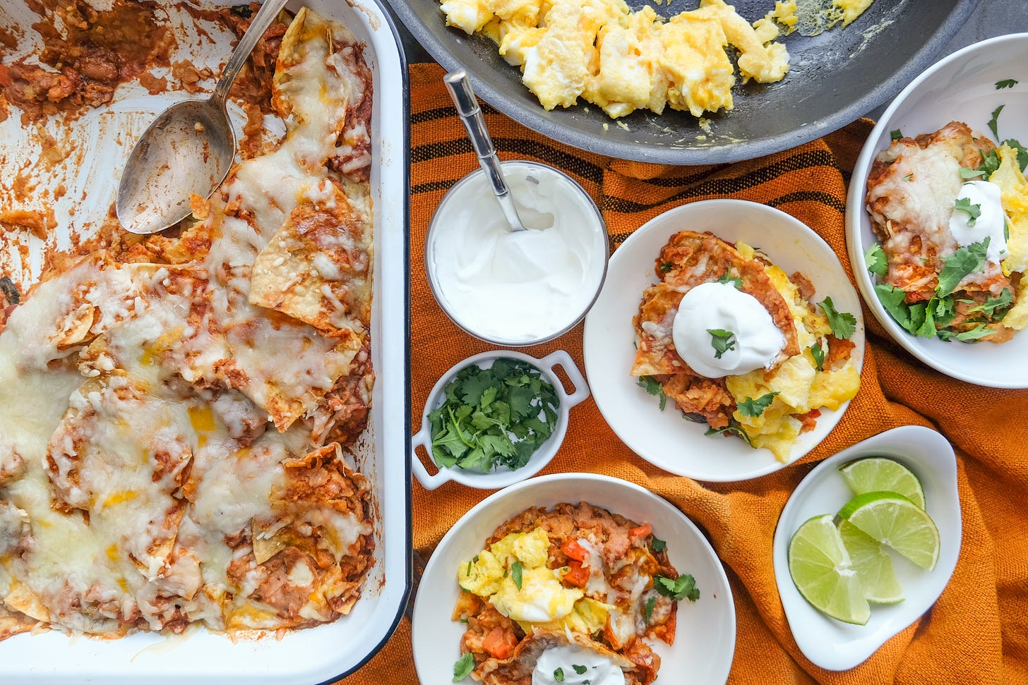 Large pan of Mexican chilaquiles, with small serving plate of chilaquiles set out next to a dish of sour cream, cilantro, and sliced lime, and a pan of scrambled eggs, placed on top of an orange tablecloth