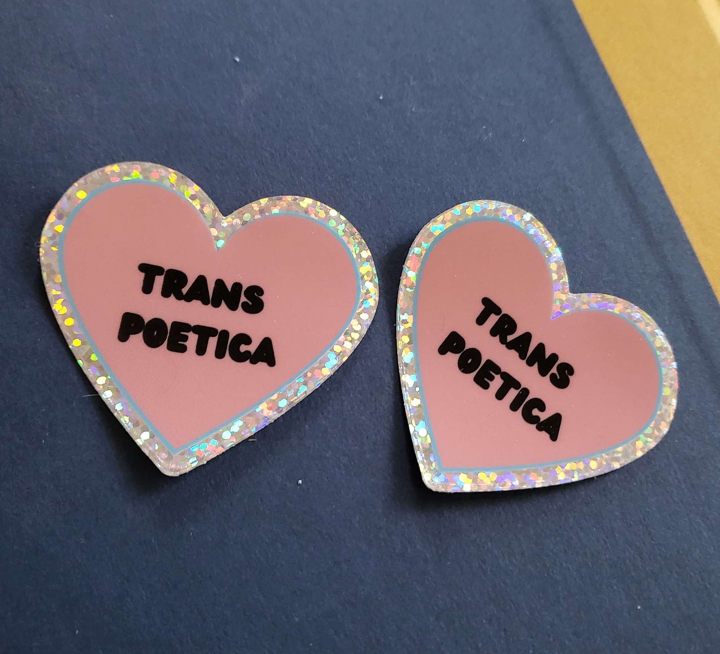 two heart shaped stickers. they are pink in the middle and say TRANS POETICA in bubble letters. there is a blue outline, and a glittery outline of the heart