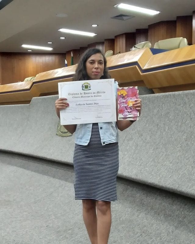 Eu, uma mulher negra de pele clara e cabelos cacheados, usando um vestido escuro com listras horizontais brancas, estou no plenário da Câmara Municipal de Goiânia segurando o mesmo diploma da foto anterior e dois exemplares da primeira edição do meu livro Ela Falam Por Si.