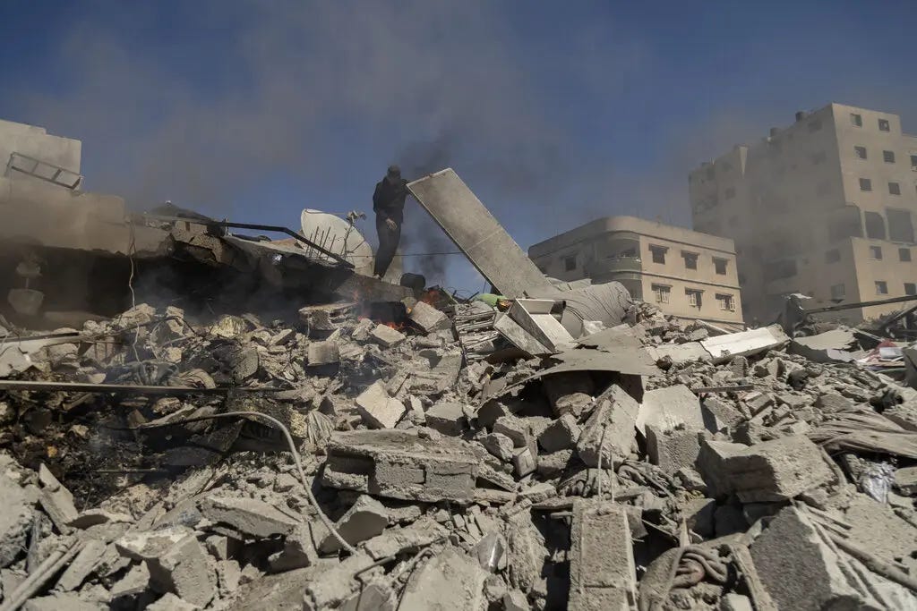 A man lifts a piece of debris atop a pile of rubble, some of it burning, that is all that remains of a building.