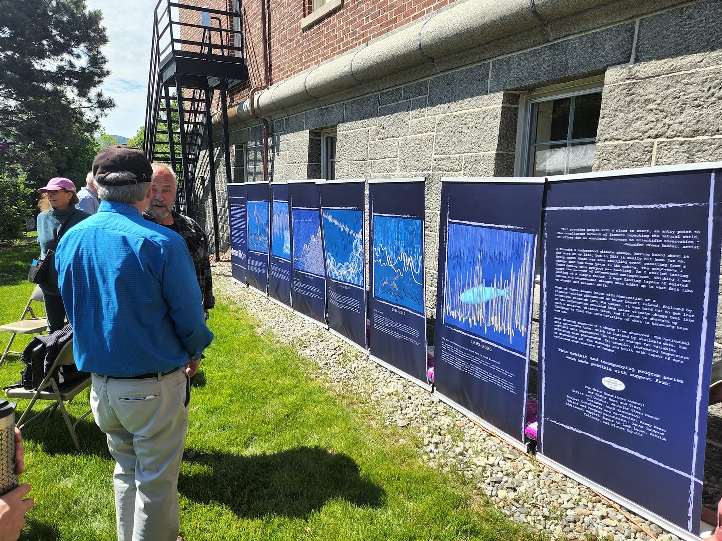 Eight large blue interpretive panels are lined up outdoors against the wall of a brick and granite block building. The panels show artwork incorporating data graphs. Four people are standing and talking nearby.