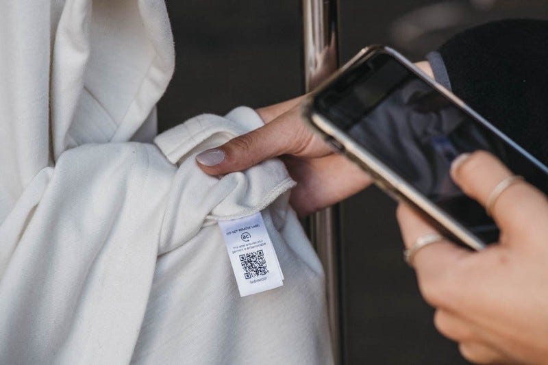 Materials science and manufacturing company Avery Dennison has partnered with textile waste recycling specialist Ambercycle to launch an intelligent digital care label solution (2021). Image shows the care label of a garment with QR code, and a person holds their phone up to the code to scan it.