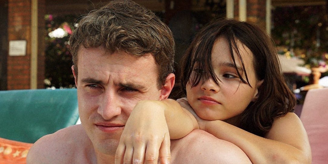 A pale white man with light brown hair looks with a furrowed brow into the distance while a young girl with dark brown hair, which is messily falling over her face, drapes her right arm over him as she looks intently at him.