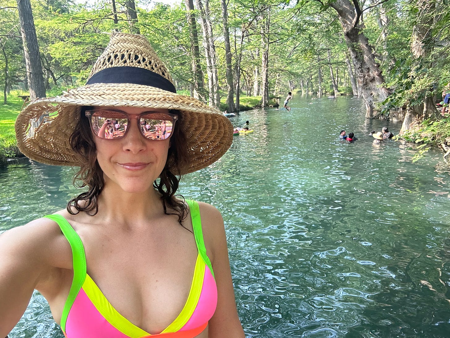 A women wearing a pink bikini top, reflective sunglasses, and a straw hat takes a selfie with a swimming hole behind her.