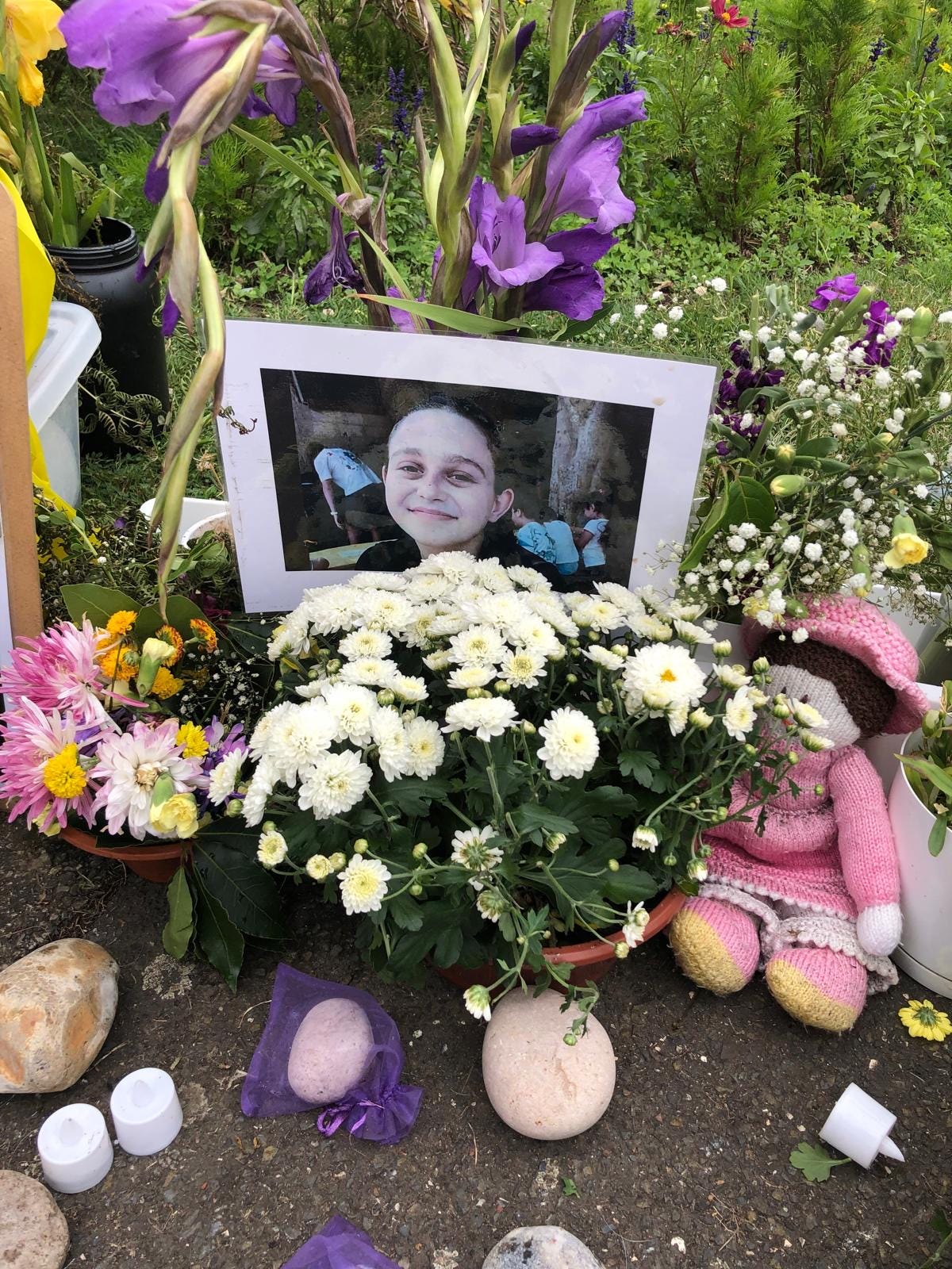 A young woman smiling in a photograph sits amongst white and yellow flowers at a memorial site