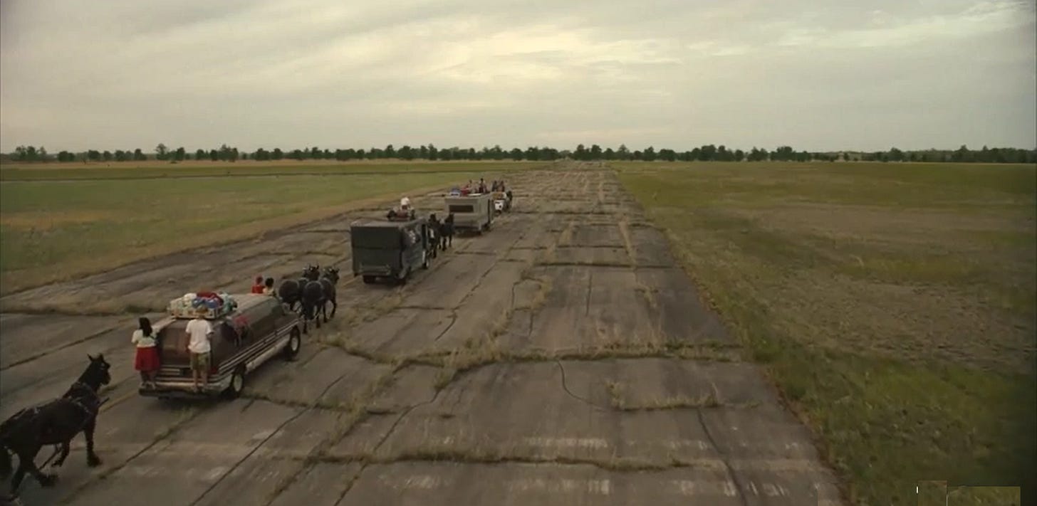 A train of vehicles drawn by horses procedes up a cracked, abandoned road toward a line of trees visible on the horizon.