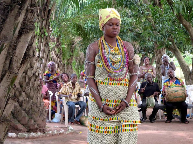 Several people play musical instruments and sing while sitting in the background while a woman adorned with numerous glass beads stands in the foreground.