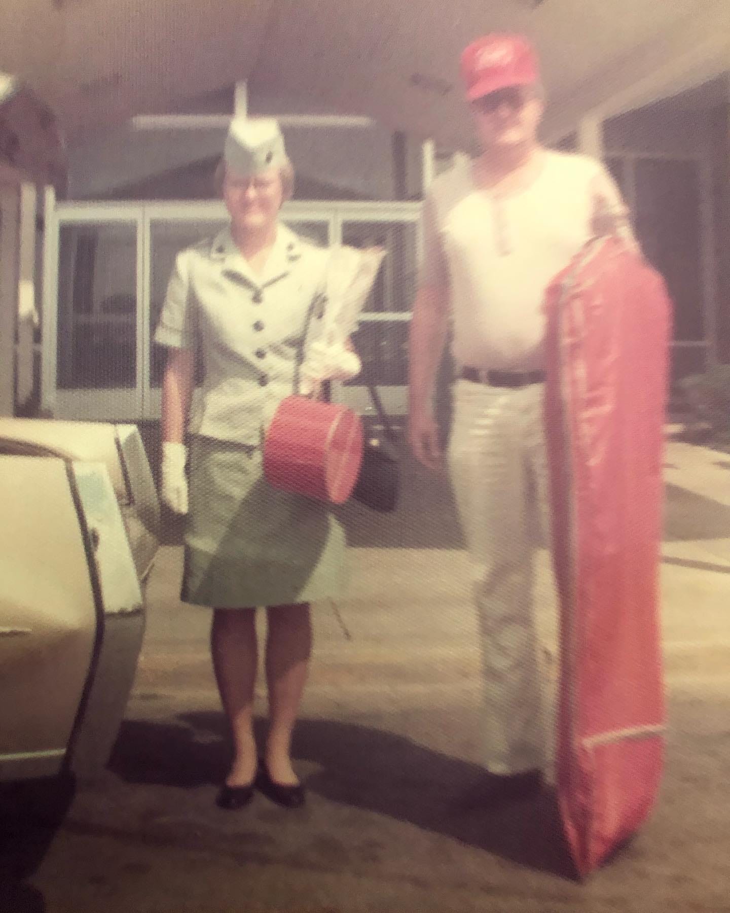A young woman in 1976 Marine Corp dress suit stands next to a man, each carrying a piece of a red luggage set
