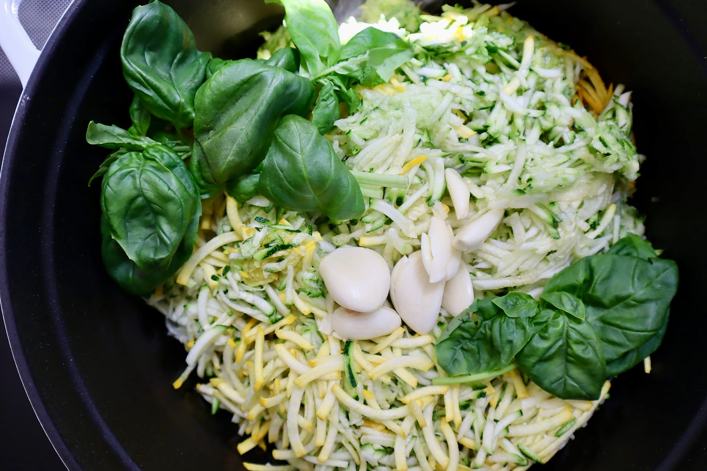 Large pot with shredded zucchini, peeled garlic cloves, and whole leaves of basil.