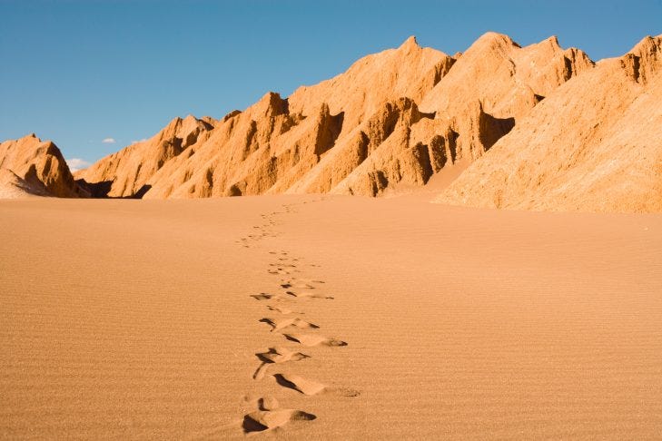 Footprints mark a great expanse of desert sand with high sandy hills running along the border under a blue sky.