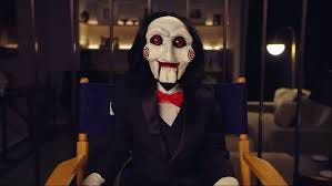 Billy the Puppet from SAW X sits in a yellow armchair