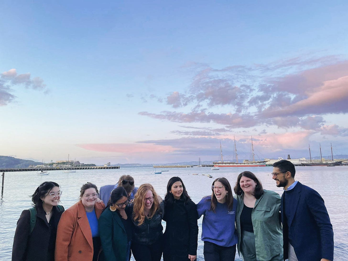 Nine people laughing with arms around each other's shoulders, standing in front of a bay at sunset