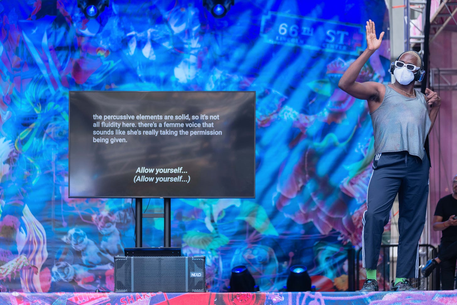 On a stage against a blue backdrop, there is a large TV screen showing two kinds of captions. One describes the aesthetics of a song: “the percussive elements are solid, so it’s not all fluidity here. there’s a femme voice that sounds like she’s really taking the permission being given.” Before, captions of the lyrics: “Allow yourself… (Allow yourself…)” Next to the monitor is Jerron Herman, a dancing disabled Black man wearing chic club attire, white sunglasses, a white KN95 mask, and headphones.