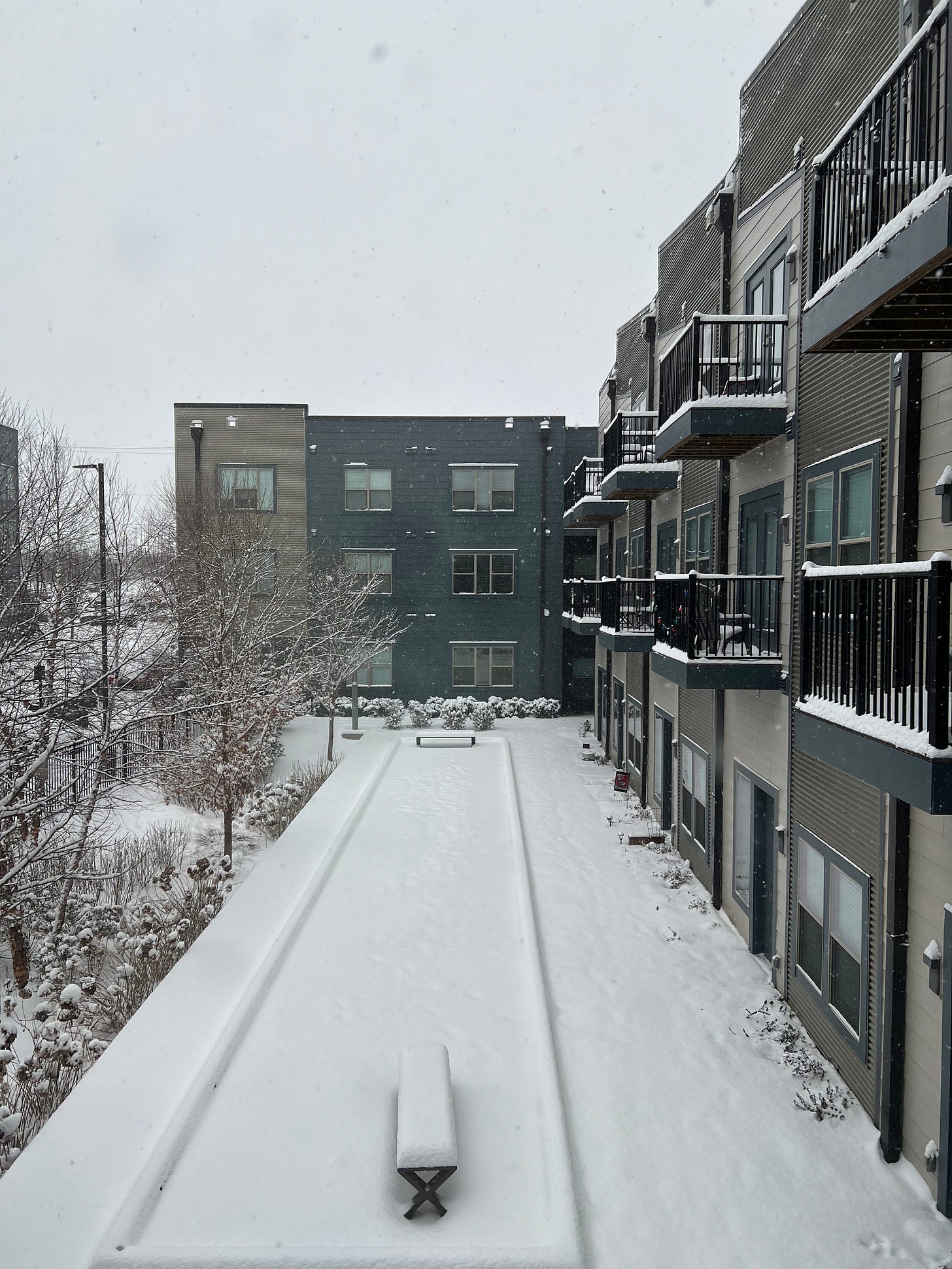 Image of snow atop a bocce ball court, many apartment patios, and barren wintry trees, at an apartment complex