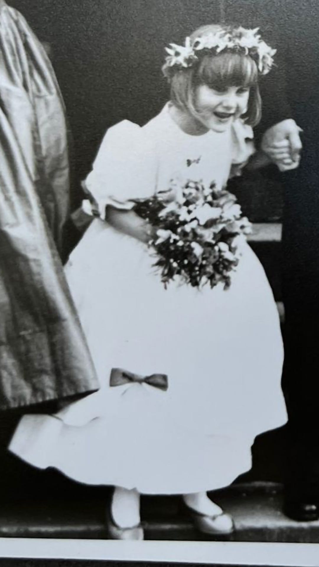 Hannah, aged five, in a black and white photo, wearing a white meringue flower girl dress with blue ribbons and blue shoes, a floral headband, and carrying a small bouquet. She is holding the hand of the groom who is out of shot due to cropping. 