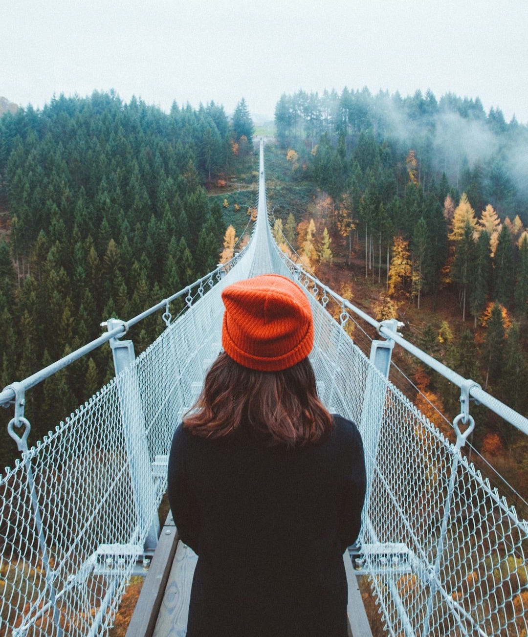 woman wearing knit cap focused on walking on white bridge between trees during daytime