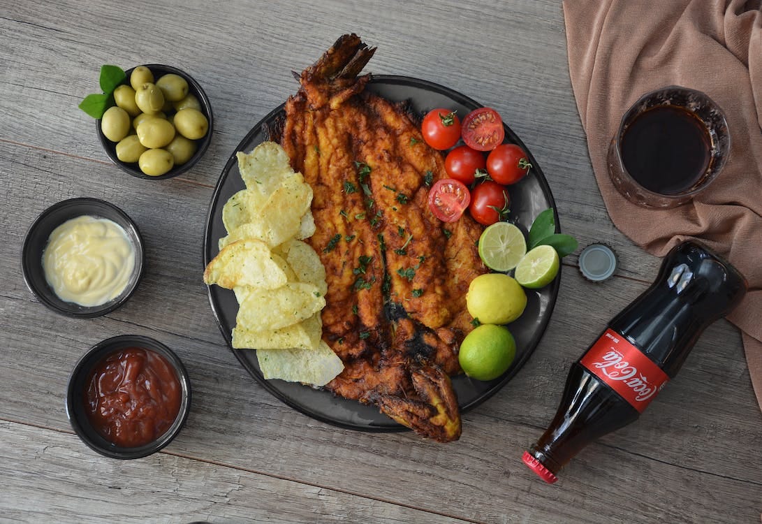 Free Top view of appetizing fried fish garnished with crunchy potato chips with limes and cherry tomatoes near ketchup and glass of refreshing drink Stock Photo