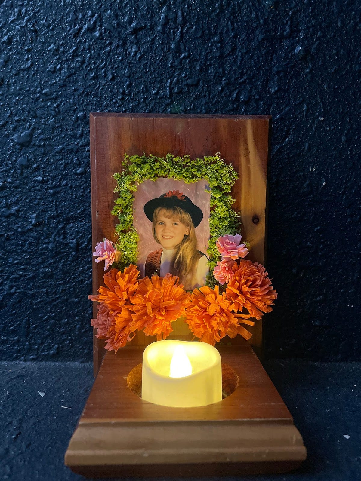 a dark blue wall background. There is a brown wooden wall sconce with a picture of a 11 year old girl with blonde hair and a black bowler hat with a red flower on it. The girl is smiling at the camera and is wearing a vest. Surrounding the picture are clumps of green moss and orangea nd pink flowers cut out of paper. There is an electric candle in front. It is an altar to the author's inner child.