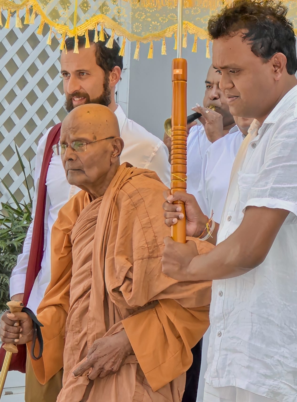 Elderly monk in saffron robes walks accompanied by attendants in white carrying and umbrella and blowing theour a horn.