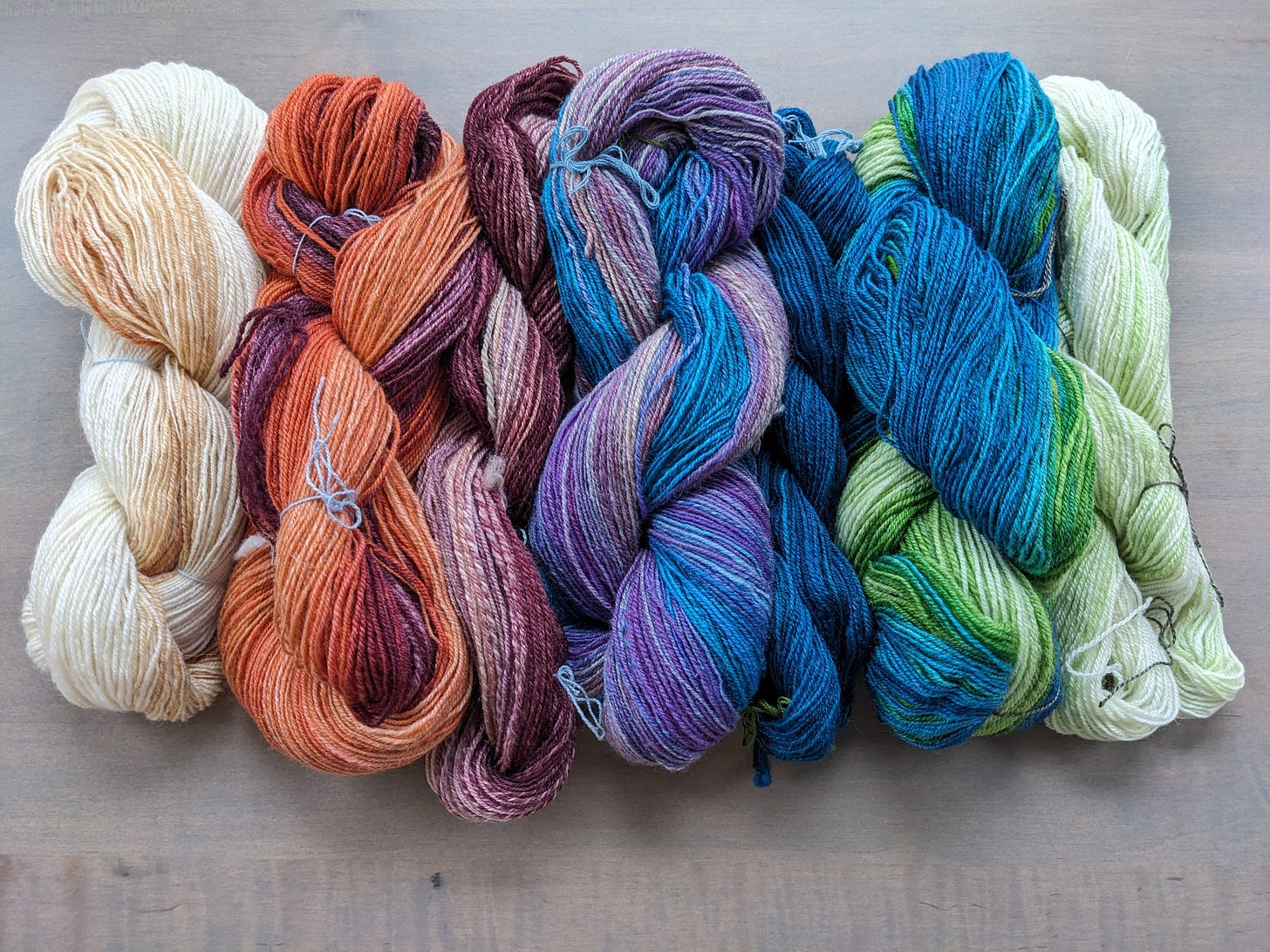 A series six skeins of yarn in a gradient from cream to orange to purple to blue to teal to green to cream.